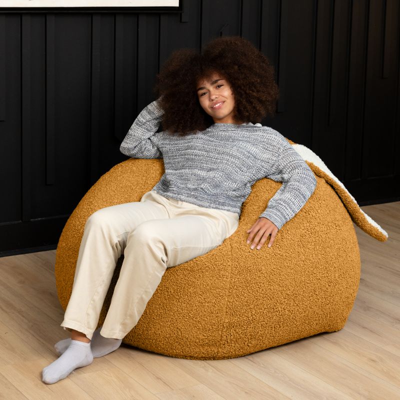 an adult reclines in the gold Raven-Symoné HopSeat sherpa bean bag chair, sinking into its deep-pressure comfort