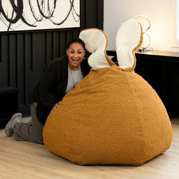 an adult playfully peeks over the gold Raven-Symoné HopSeat sherpa bean bag chair, showing its tall bunny-ear design