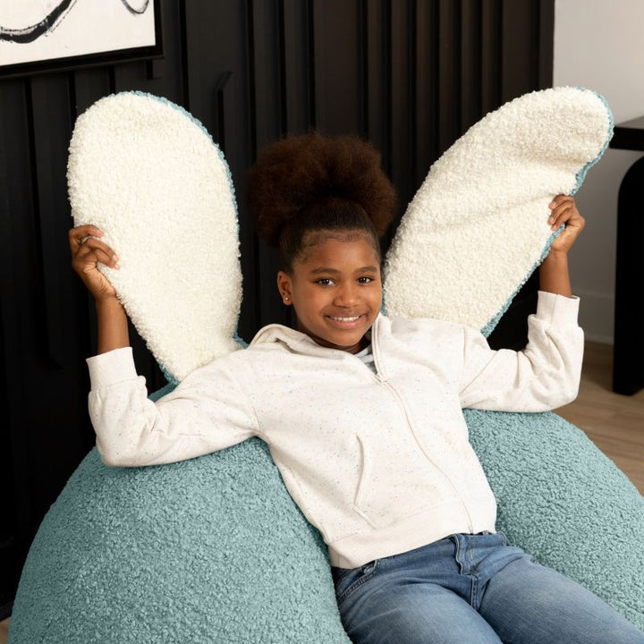 a child sits in the green Raven-Symoné HopSeat sherpa bean bag chair while holding up its soft, tactile bunny ears