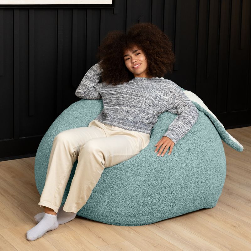 an adult reclines in the green Raven-Symoné HopSeat sherpa bean bag chair, sinking into its deep-pressure comfort