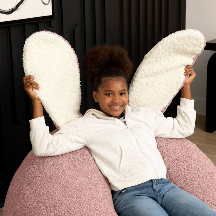 a child sits in the pink Raven-Symoné HopSeat sherpa bean bag chair while holding up its soft, tactile bunny ears
