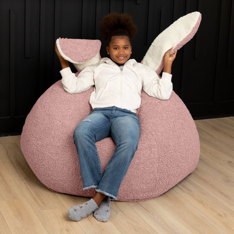 a child sits in the pink Raven-Symoné HopSeat sherpa bean bag chair while holding up its soft, tactile bunny ears