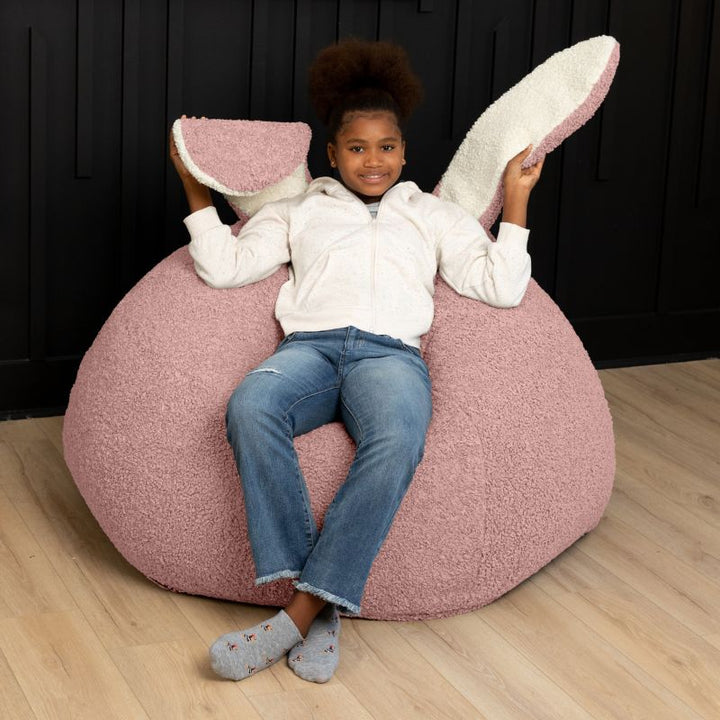 a child sits in the pink Raven-Symoné HopSeat sherpa bean bag chair while holding up its soft, tactile bunny ears