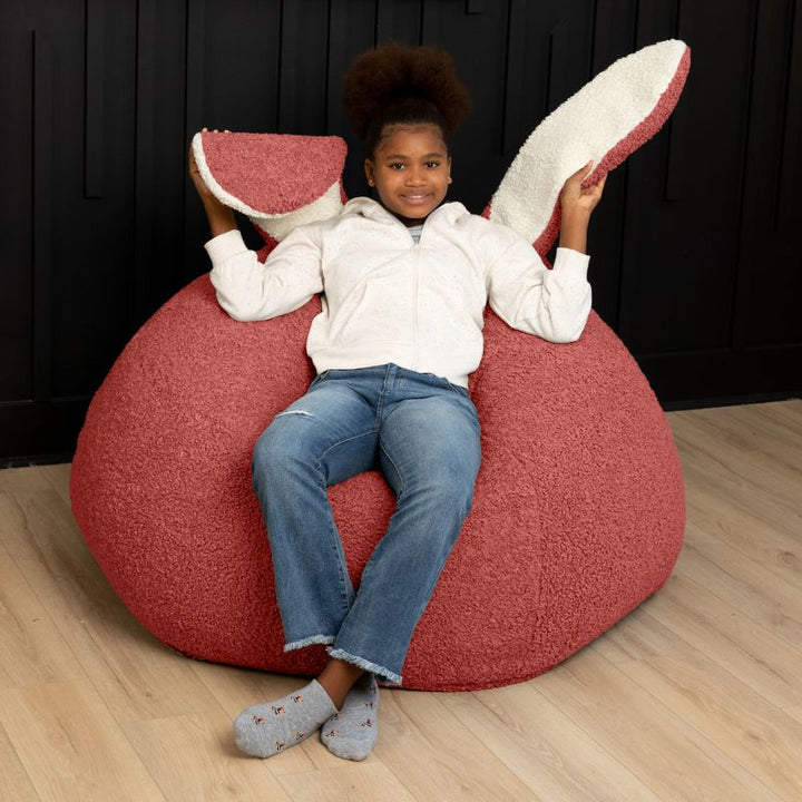 a child sits in the Rosewood Red Raven-Symoné HopSeat sherpa bean bag chair while holding up its soft, tactile bunny ears