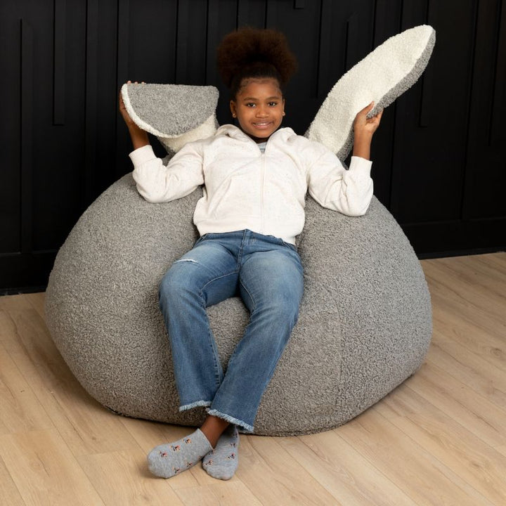 a child sits in the Smoke Raven-Symoné HopSeat sherpa bean bag chair while holding up its soft, tactile bunny ears