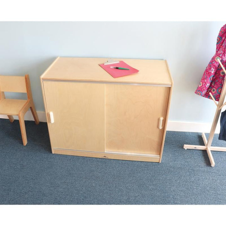 Whitney Brothers Sliding Doors Storage Cabinet with doors closed in a bright classroom, a birch plywood classroom storage cabinet beside a child's chair and coat tree