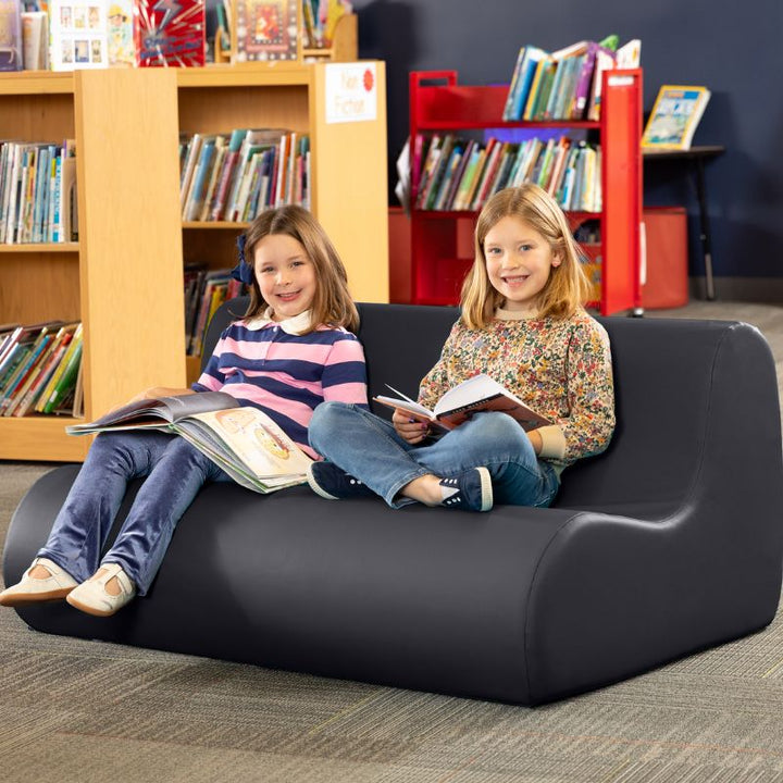 Two children reading on a black Jaxx Midtown soft seating couch in a school library calm corner