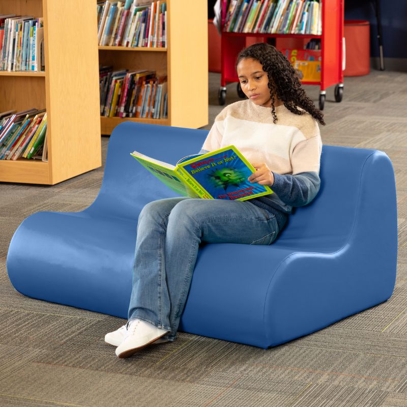 A student reading on a blue Jaxx Midtown soft seating couch in a school library setting