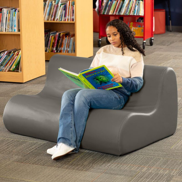 A student reading on a charcoal Jaxx Midtown soft seating couch in a school library setting