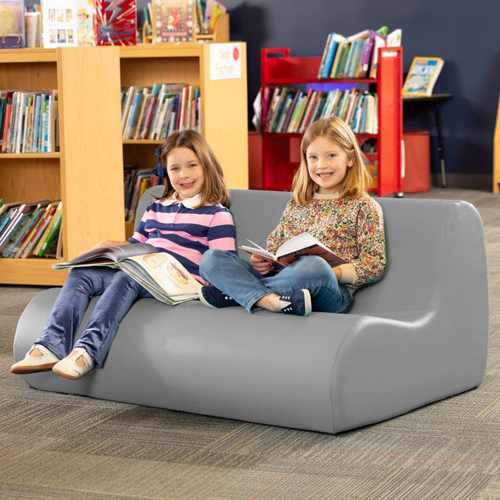 Two children reading on a grey Jaxx Midtown soft seating couch in a school library calm corner