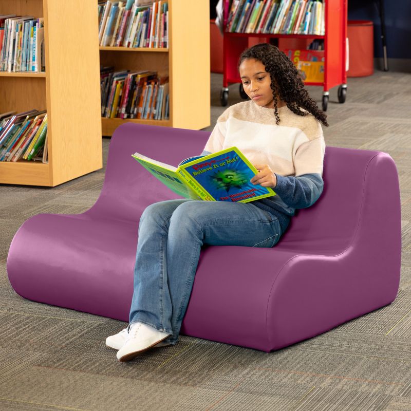 A student reading on a purple Jaxx Midtown soft seating couch in a school library setting