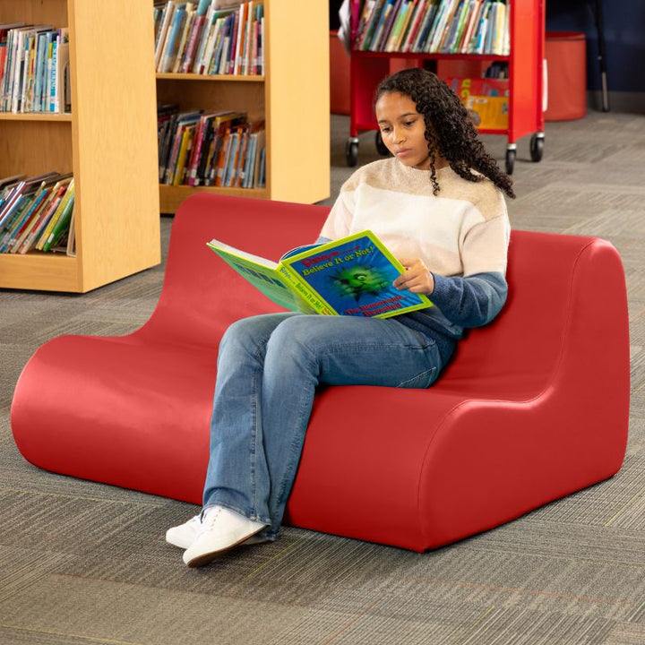 A student reading on a red Jaxx Midtown soft seating couch in a school library setting