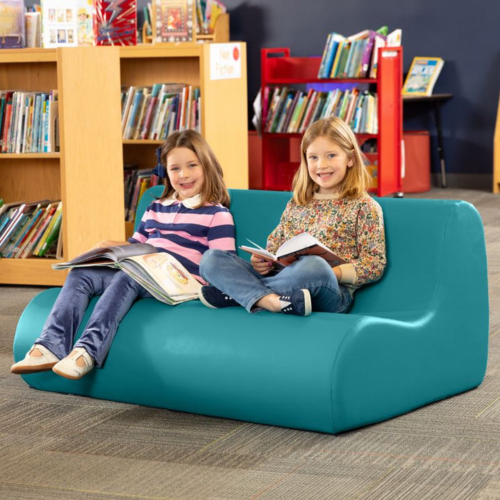 Two children reading on a teal Jaxx Midtown soft seating couch in a school library calm corner