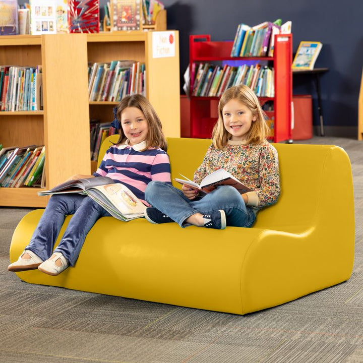 Two children reading on a yellow Jaxx Midtown soft seating couch in a school library calm corner