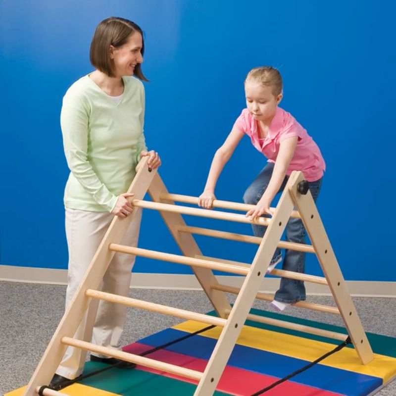 Child climbing over the top of the Southpaw Angle Ladder, a wooden A-frame climbing ladder set up on a tumbling mat as part of a sensory obstacle course, with an adult spotting alongside