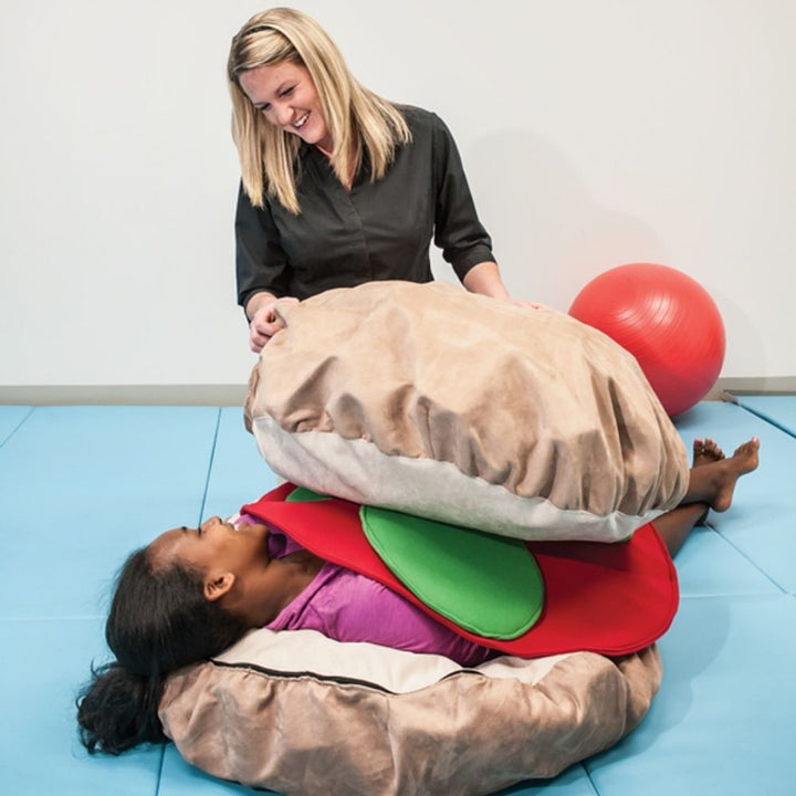 Southpaw Weighted Hamburger with adult woman and child lying on the plush floor pillow bun, demonstrating whole-body deep pressure proprioceptive input