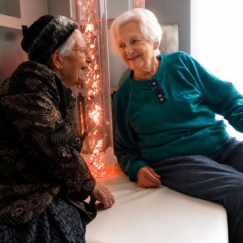 Two older women smiling and chatting beside the TFH Aquarium Bubble Tube 6" glowing orange, a calming LED bubble column for care homes and senior sensory rooms