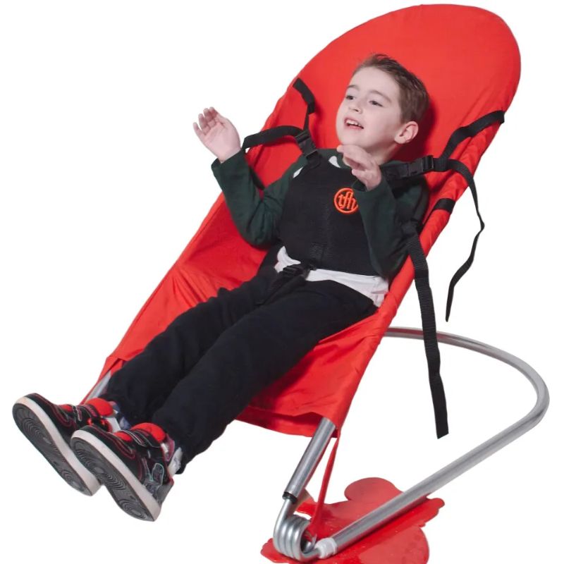 Smiling child bouncing in the TFH Bouncing Chair large red variant with the deluxe harness secured, an active vestibular sensory seat for movement-based play