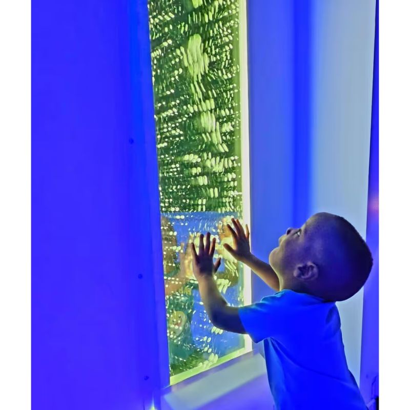 A boy reaches up to touch a TFH Bubble Wall lit in blue-green light, a sensory bubble wall for tactile and visual play