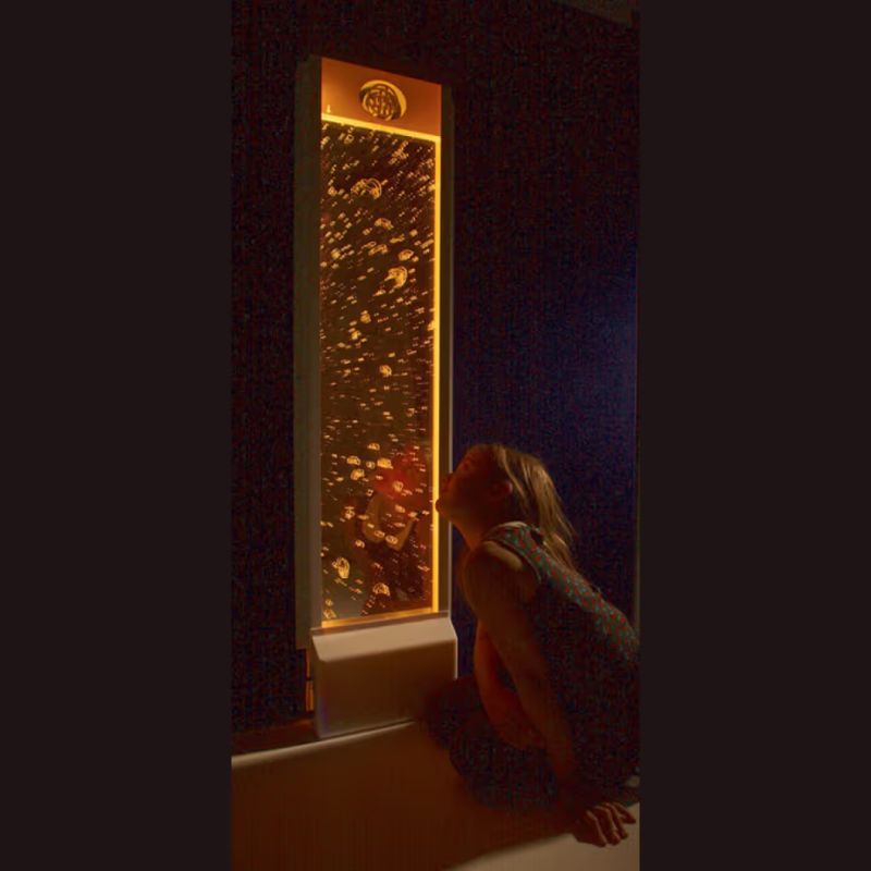 A girl looks closely at a TFH Bubble Wall lit in warm amber light, a sensory bubble wall for visual tracking and calm