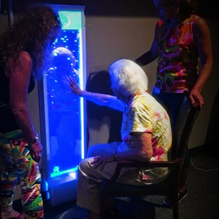 An older woman touches a TFH Bubble Wall lit in blue light while caregivers look on in a therapy setting