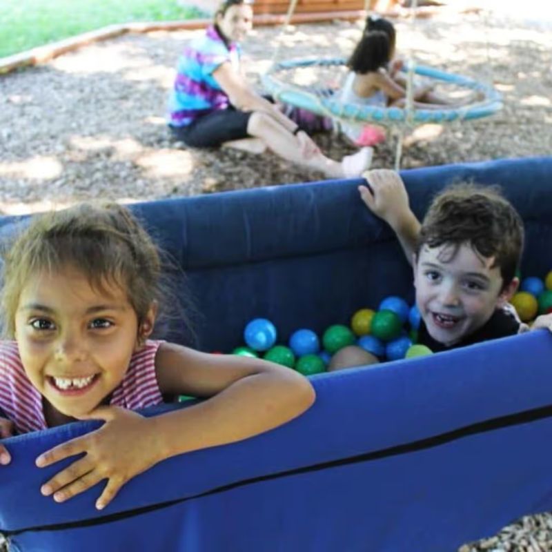 Two children playing together in the TFH Deep Boat Swing outdoors, an enclosed sensory swing offering shared vestibular and tactile play