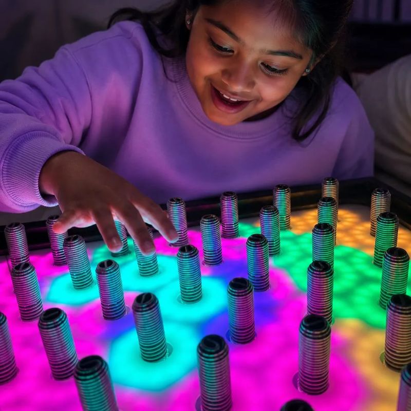 Smiling girl reaching across the TFH Electro Wobble Garden Activity Panel as pink, teal and green light blooms beneath the springs she touches