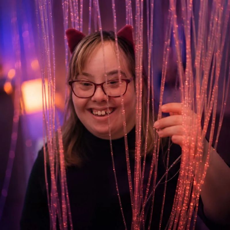 Smiling young woman peeking through the pink-lit tails of the TFH Fibre Optic Waterfall, a ceiling-suspended fibre optic sensory light for all ages and abilities