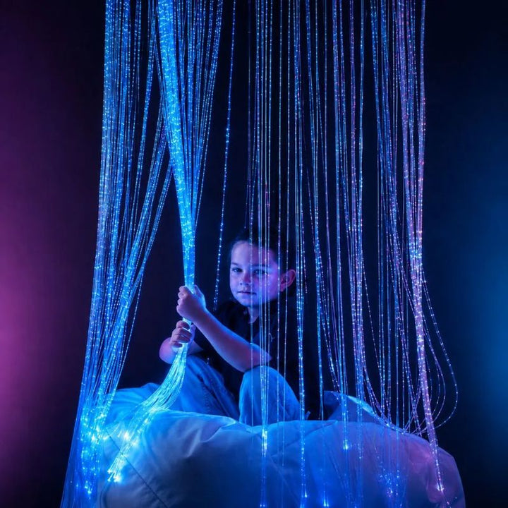Child sitting on a bean bag beneath the TFH Fibre Optic Waterfall, holding a bundle of glowing blue fibre optic tails for calming tactile and visual exploration