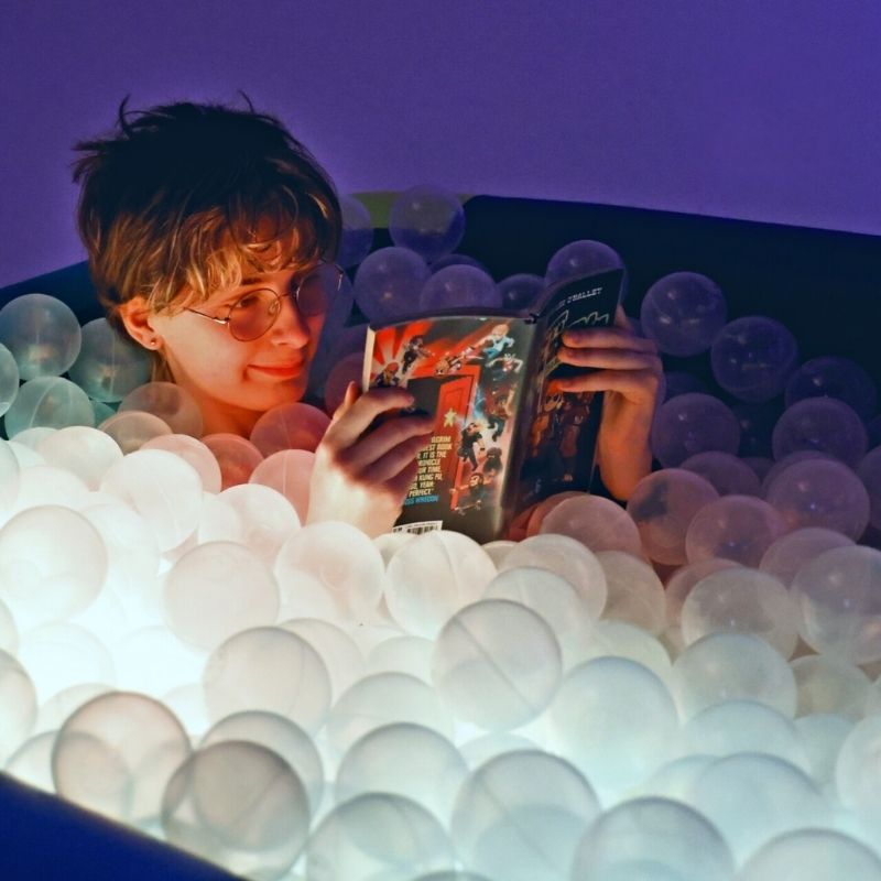 Child actively playing and reading inside the TFH Genie Illuminated Ball Pool, surrounded by 1,500 clear LED-lit balls for proprioceptive sensory play and visual stimulation