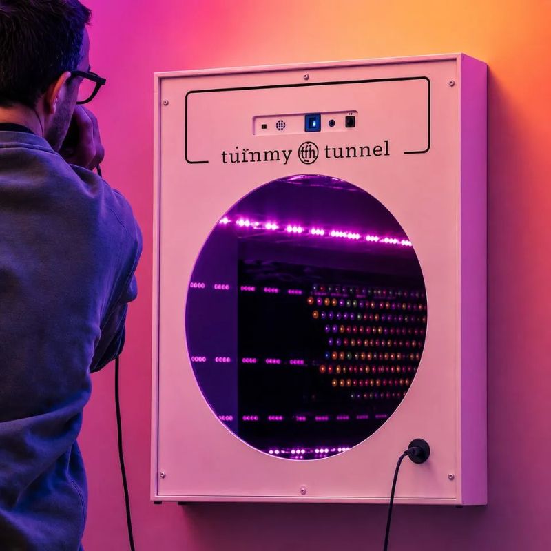 Man speaking into the microphone beside the wall-mounted TFH Interactive Infinity Tunnel, purple and multicolour LED rows glowing in its infinite-depth illusion