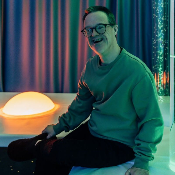 Adult sitting beside the TFH Interactive Dome Swirl Podium, showing the accessible eye-level height and personal engagement the podium mount creates for sensory room users