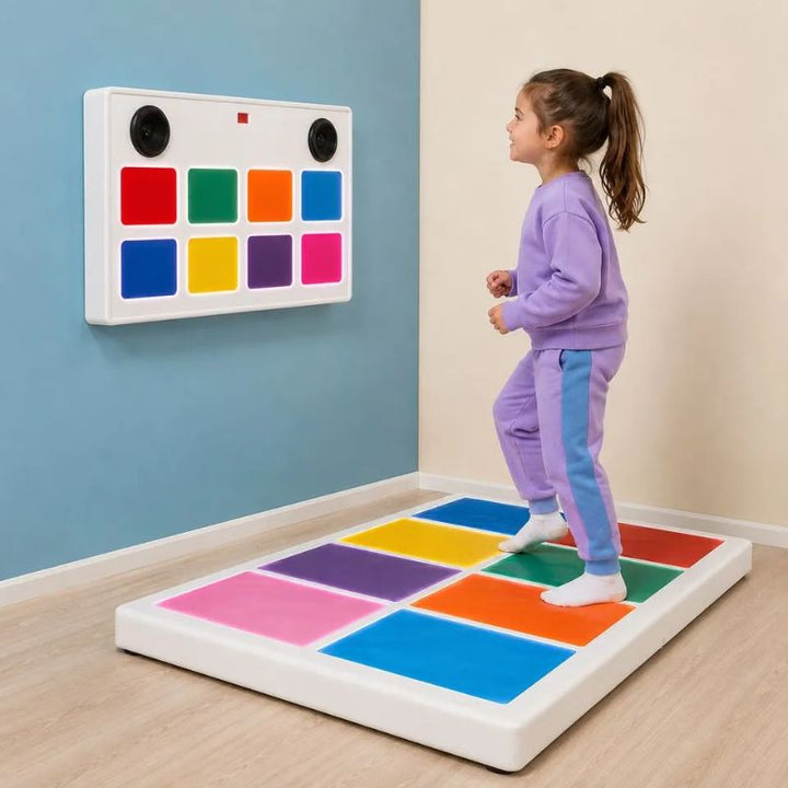 Smiling girl stepping across the colour squares of the TFH Interactive Musical Squares floor pad, with the 8-switch musical panel mounted on the wall behind her