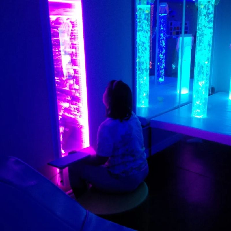 Child sitting close to watch the TFH Lights & Bubbles Water Panel glowing purple in a multi-sensory room, a space-saving bubble wall for visual tracking and calm