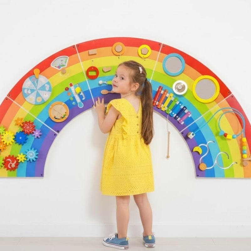 Child engaged with the TFH Rainbow Wall Activity Panel, showing the full arc installation with 5 colourful panels and 12 hands-on sensory activities for early learning