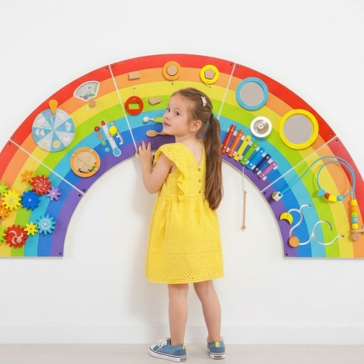Child engaged with the TFH Rainbow Wall Activity Panel, showing the full arc installation with 5 colourful panels and 12 hands-on sensory activities for early learning