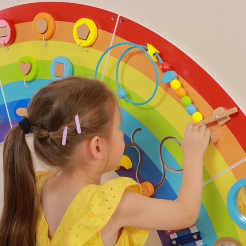 Close-up of a child interacting with the TFH Rainbow Wall Activity Panel, showing fine motor activities and the vibrant rainbow-themed sensory busy board design
