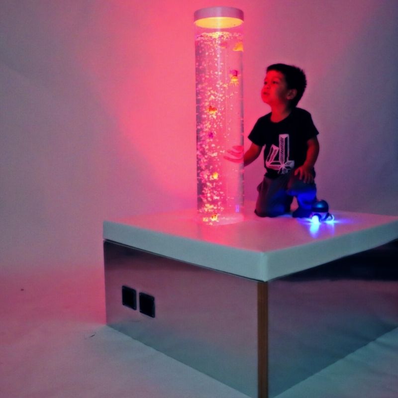 TFH Square Podium Base in a sensory room landscape view with a child actively playing beside the bubble tube, showing the padded mirrored square podium for close-contact sensory engagement