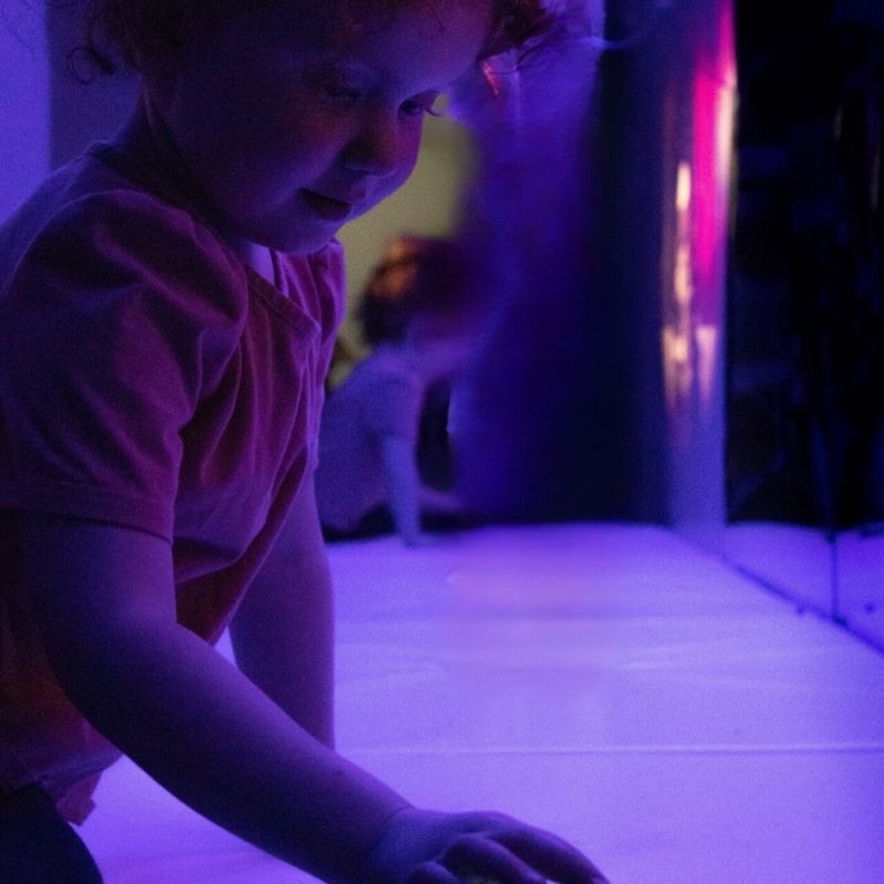 Close-up of the TFH Square Podium Base with a child actively engaging with the bubble tube, showing the padded vinyl seating surface for close-contact sensory play in a therapy room