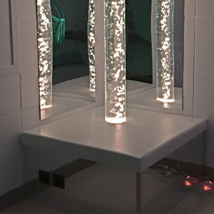 TFH Square Podium Base in a sensory room landscape view with the bubble tube glowing in warm light, showing the mirrored sides reflecting colour across the sensory room floor