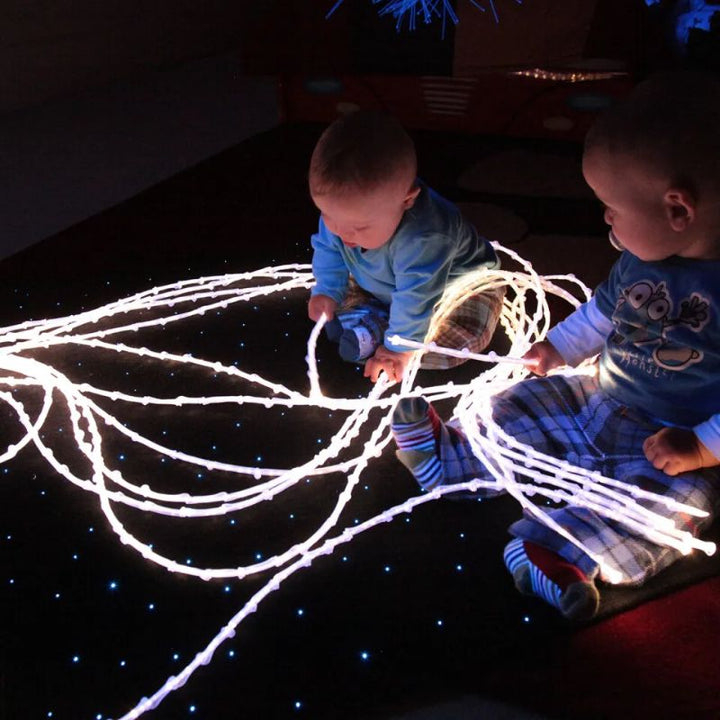 Two babies exploring the glowing knotted tails of the TFH Textured Light Spray on a sparkling floor, a tactile fibre optic spray for early sensory play