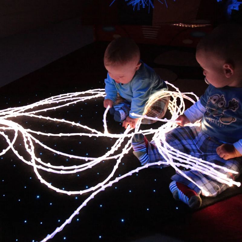 Two babies exploring the glowing knotted tails of the TFH Textured Light Spray on a sparkling floor, a tactile fibre optic spray for early sensory play