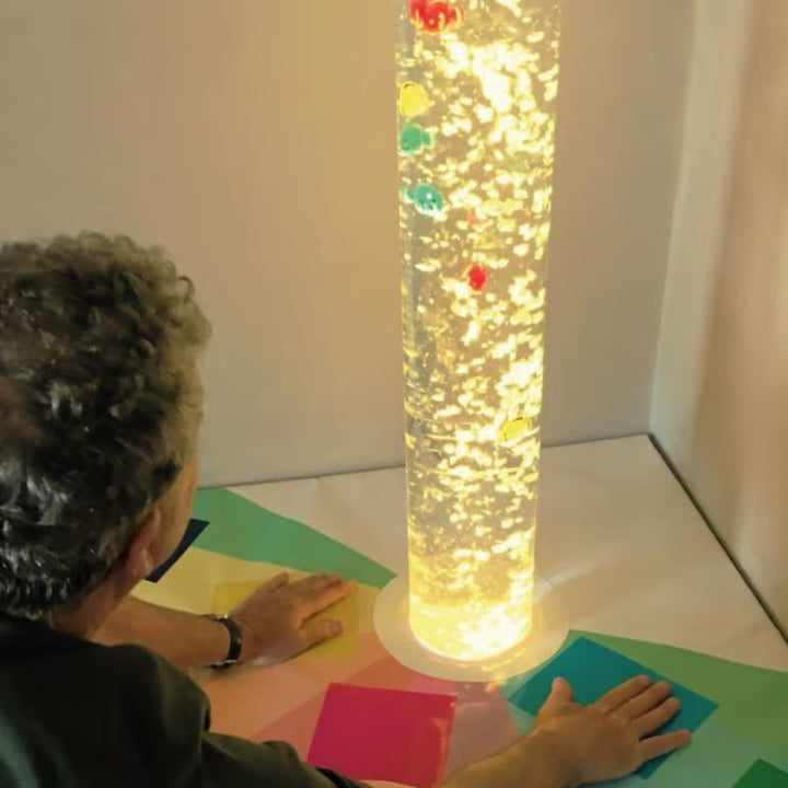 An adult touches the TFH Touch-A-Color Podium pad beside a bubble tube with a floating fish insert for extra visual stimulation
