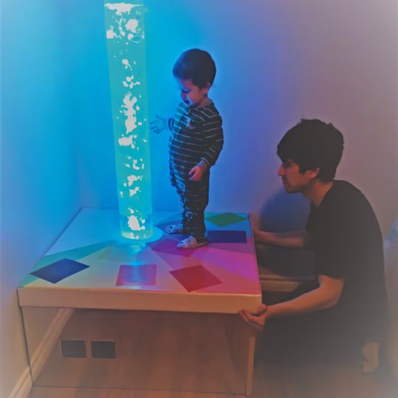 A child and caregiver interact with the TFH Touch-A-Color Podium as the bubble tube glows teal in a multi-sensory room