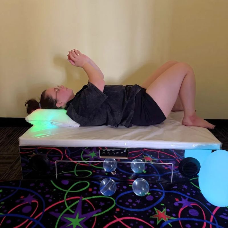 Adult relaxing on the TFH Vibroacoustic Platform with Foam Pad in a multi-sensory room, feeling low-frequency sound vibration for calm and sensory regulation
