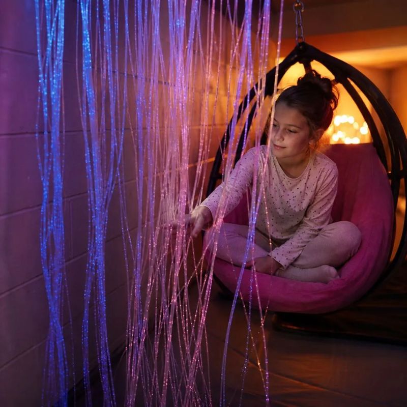 Girl relaxing in a hanging egg chair as she reaches into the pink and blue strands of the TFH Wall Mounted Fiber Optic Curtain, enjoying calming tactile and visual input