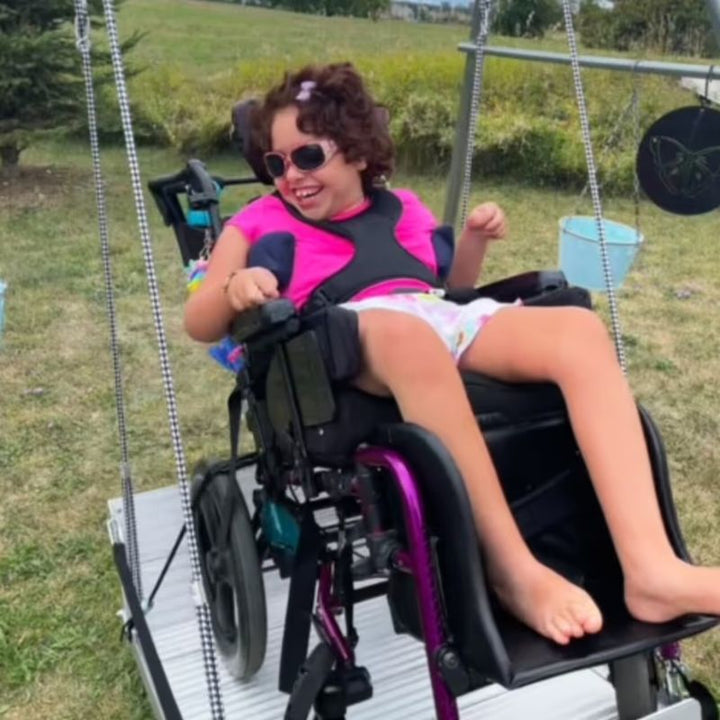 A young girl in her wheelchair enjoying the TFH Wheelchair Platform Swing outdoors, an accessible vestibular swing for wheelchair users