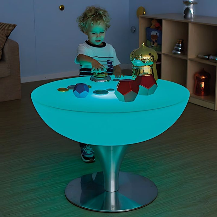 TTS Round Glow Table glowing in teal with a child reaching across the illuminated surface, showing the LED colour-changing feature in a home sensory room setting