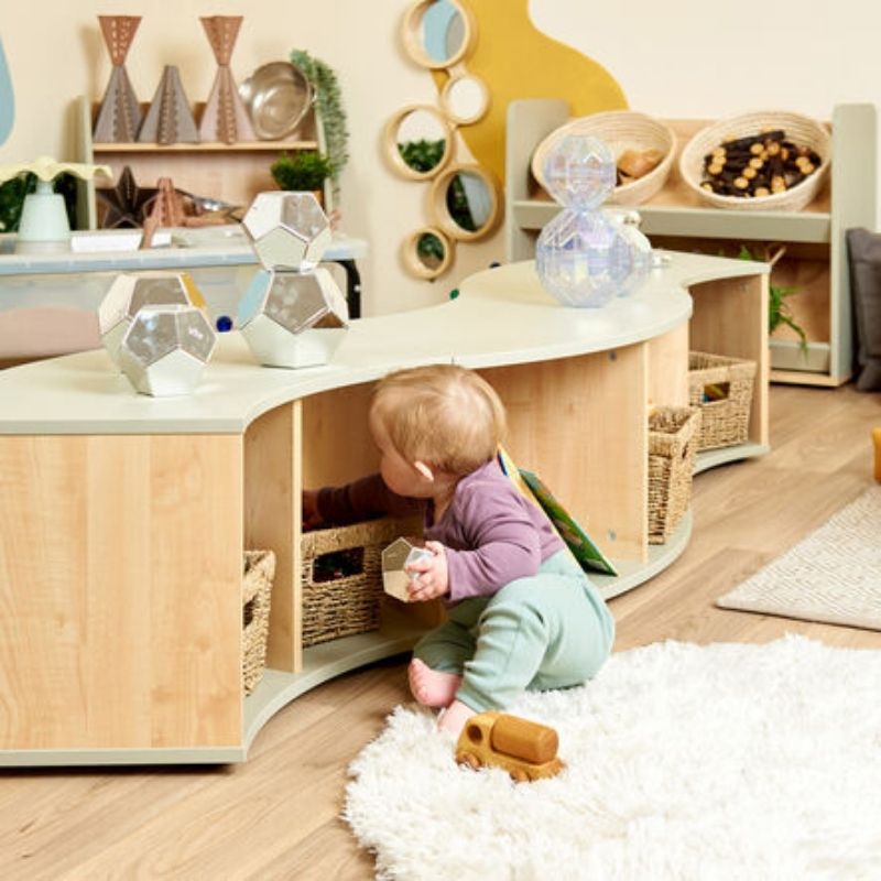Toddler sitting and playing beside the TTS Elements Sage Curved Storage Unit demonstrating gross motor engagement and independence