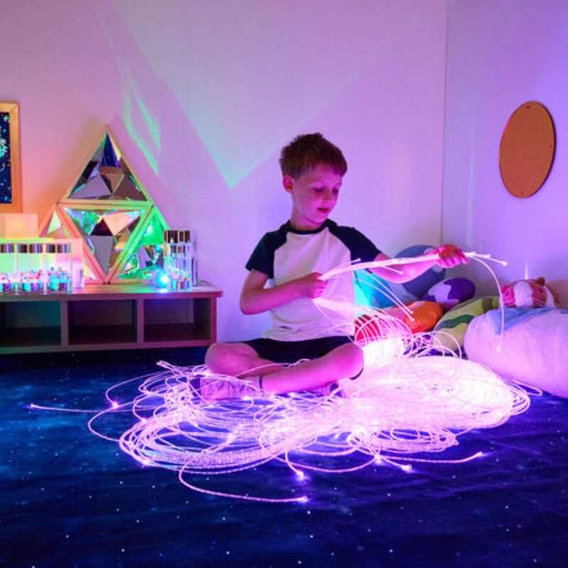 Boy sitting cross-legged lifting a bundle of glowing TTS Fibre Optic Tails with both hands, building fine motor skills in a colourful sensory room